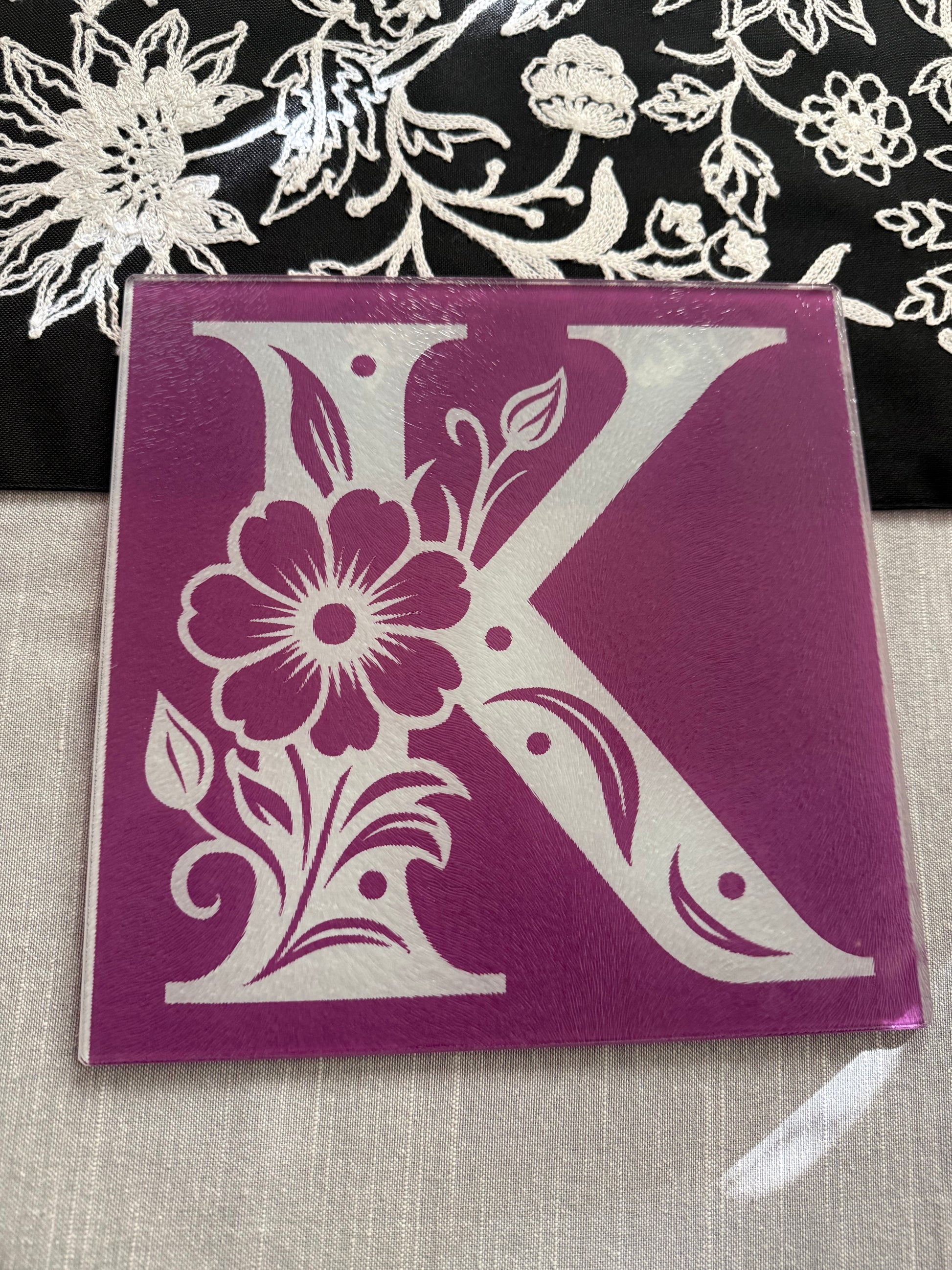 Square glass cutting board with white floral "K" design on purple background, styled with embroidered fabric, Roxzell