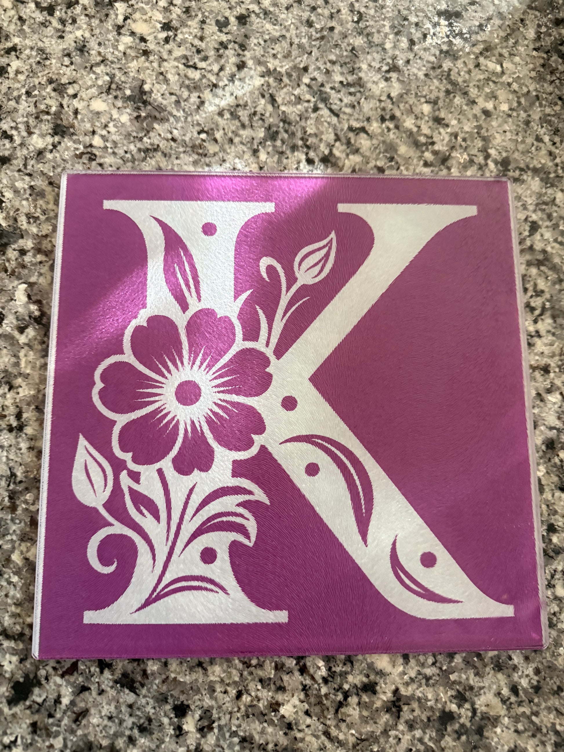 Decorative glass cutting board featuring a white floral "K" with a purple background on a granite counter, ideal for kitchen decor, Roxzell