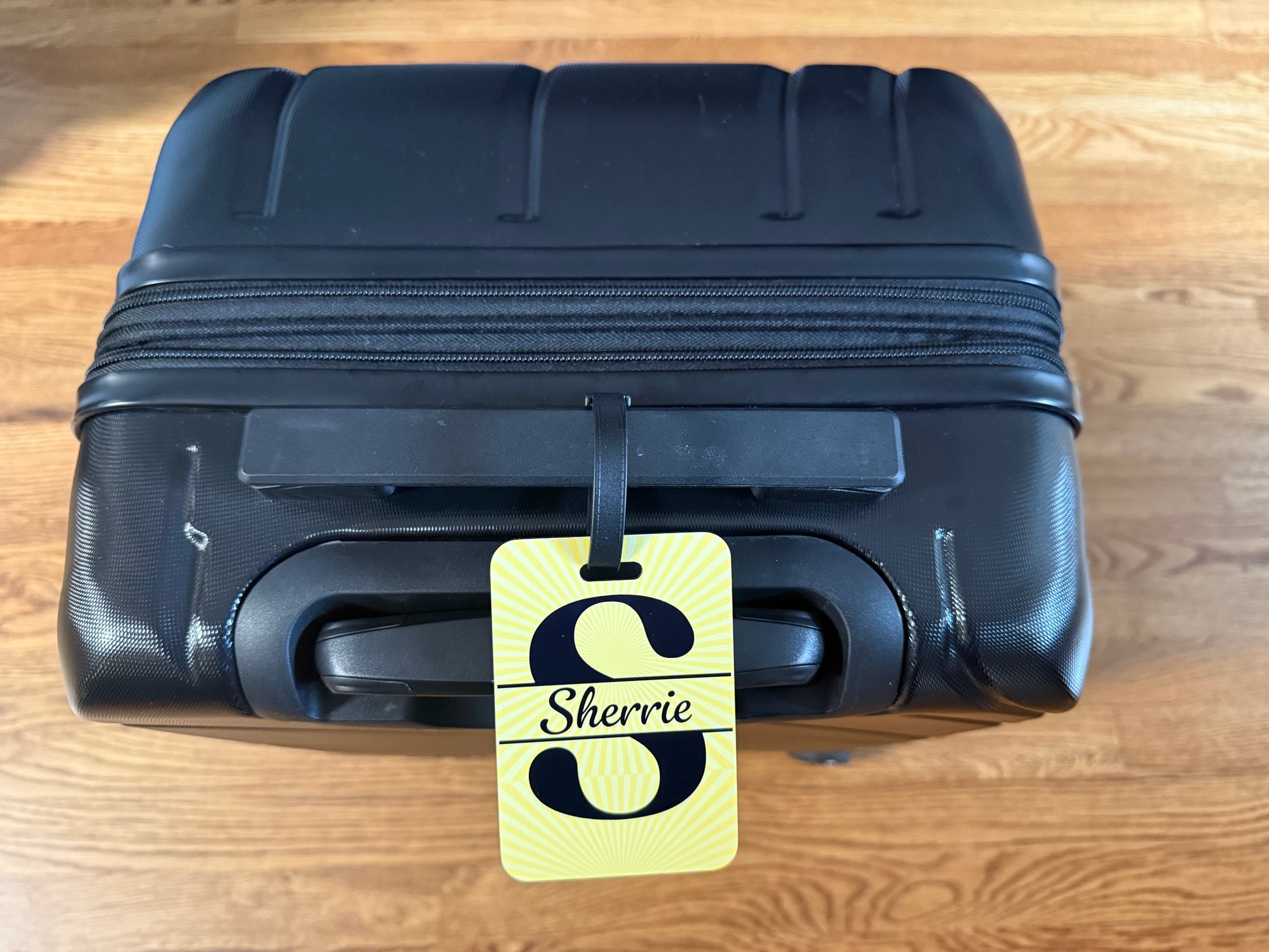 Black suitcase with a yellow luggage tag labeled 'Sherrie' on a wooden surface, Roxzell