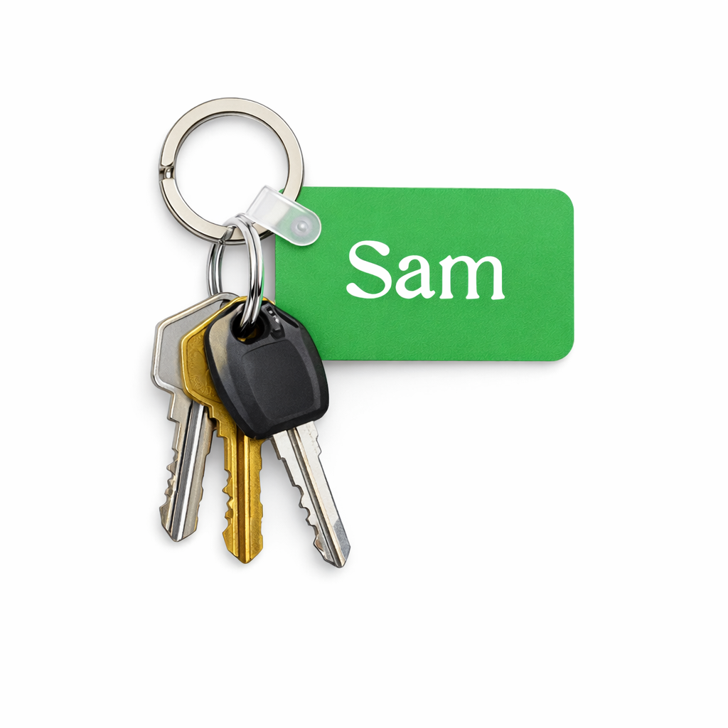 Green rectangular keychain with the name “Sam” printed vertically in white, attached to a silver ring holding three keys—one silver, one brass, and one black car key—hanging downward against a bright white background. Roxzell