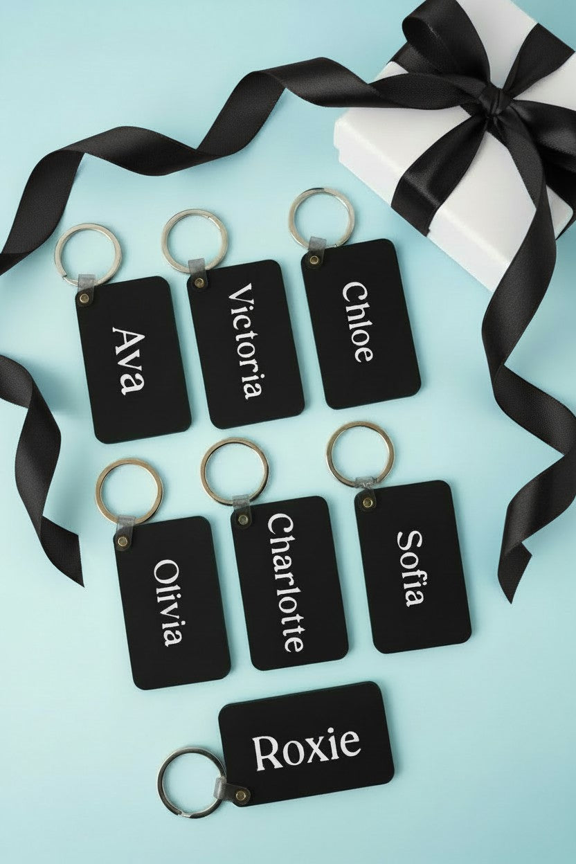 Six personalized black rectangular keychains with silver keyrings, each printed with a different name in white text, arranged on a light blue background with a white gift box and black ribbon. Roxzell