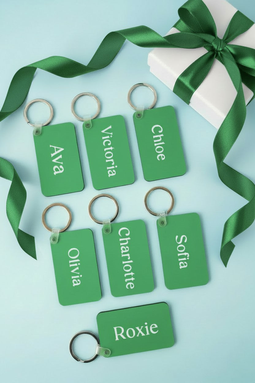 Eight personalized green gift tags with silver key rings, each printed with a different name in white text, arranged on a light blue background with a green ribbon and a white gift box. Roxzell