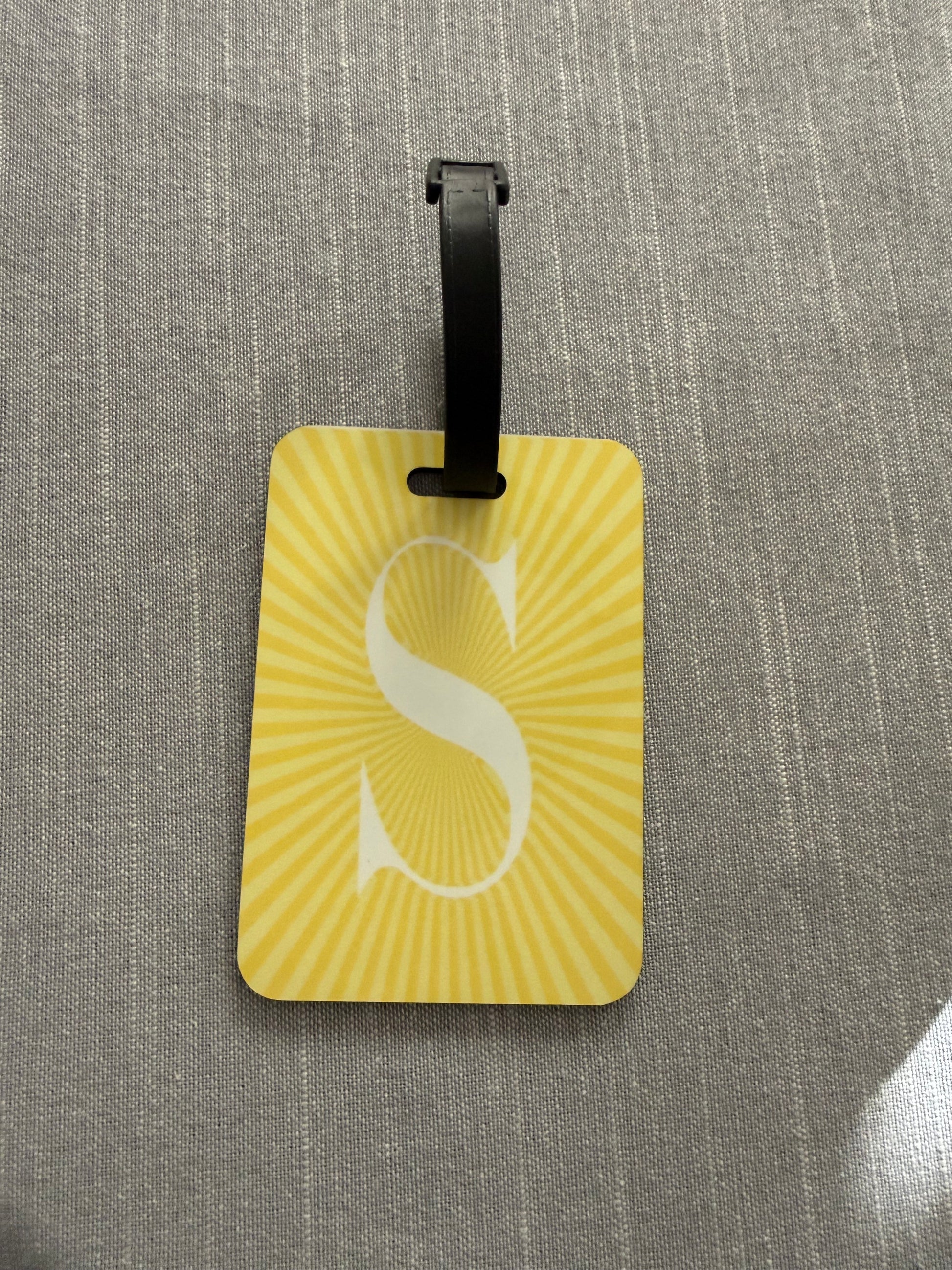 Yellow sunburst luggage tag with a black strap and white letter "S" on a gray textured surface, Roxzell