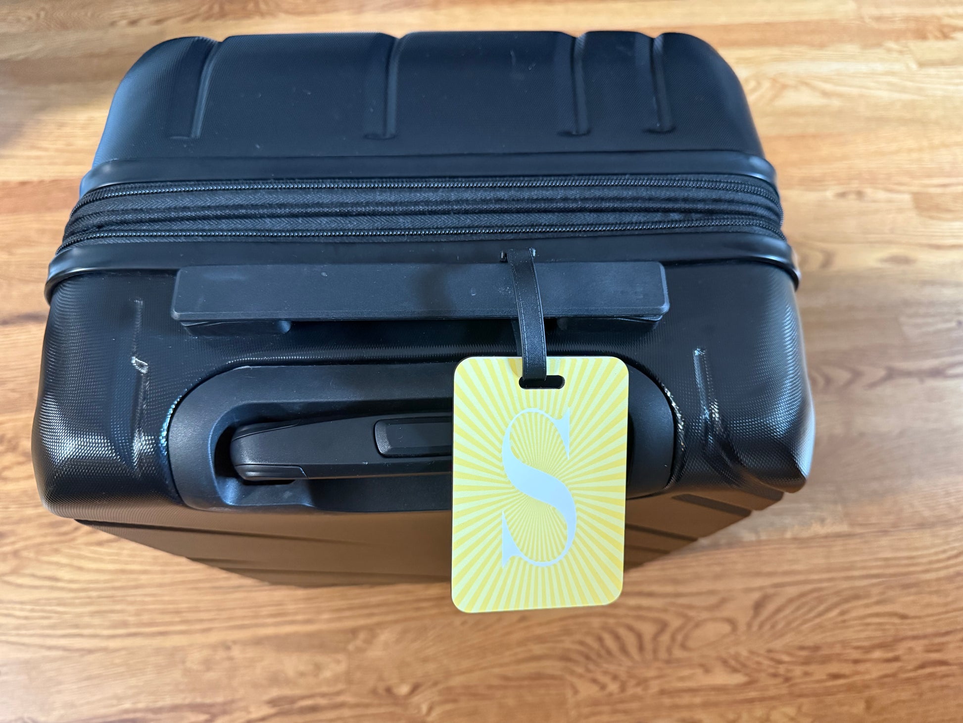Yellow luggage tag with white letter "S" and sunburst background on black suitcase, Roxzell